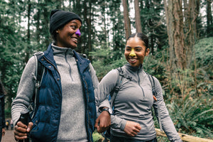 Two female hikers link arms and smile while wearing Nözscreen, our colorful, eco friendly sunscreen, in purple and yellow.